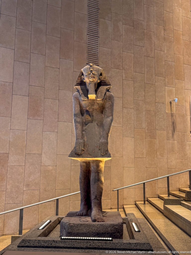 Standing ancient Egyptian royal figure with broad headdress and arms crossed over the chest, lit against a stone wall.