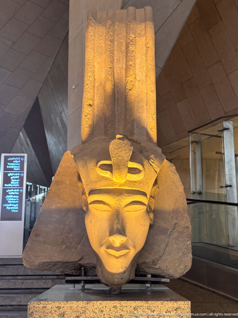 Large ancient Egyptian royal head fragment with a tall surviving headdress element, dramatically lit on a museum platform.
