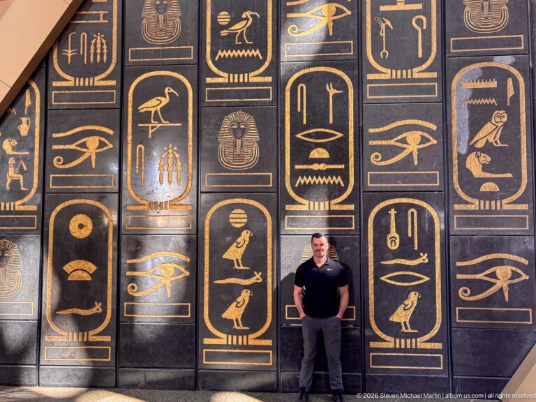 Visitor standing before tall black wall panels decorated with large gold-colored hieroglyph-inspired Egyptian motifs.