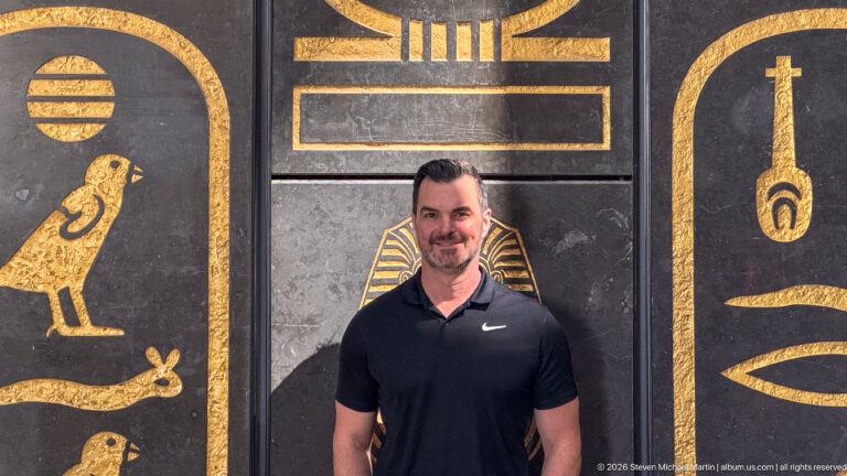 Visitor standing before black wall panels decorated with large gold-colored Egyptian birds, an eye, and hieroglyph-inspired signs.