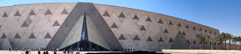 Grand Egyptian Museum exterior with pale faceted walls, a deep triangular entrance, palm trees, and visitors across the forecourt.
