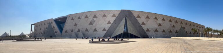 Wide exterior view of the Grand Egyptian Museum’s pale faceted walls, triangular openings, and expansive paved forecourt.
