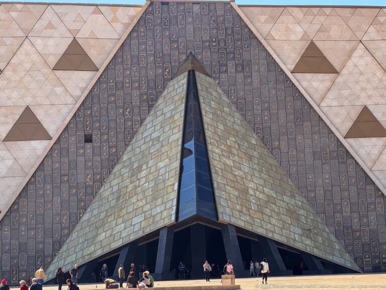 Grand Egyptian Museum façade with a deep triangular entrance recess, patterned dark panels, pale stone cladding, and visitors below.