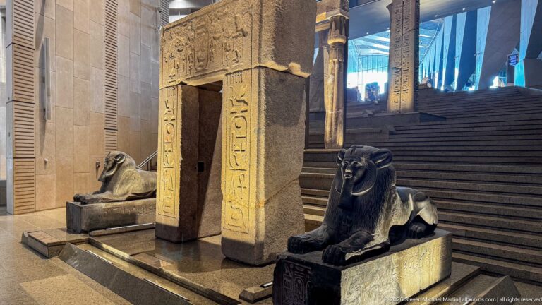 Small hieroglyph-covered stone gateway flanked by two recumbent lion statues on a Grand Staircase platform.