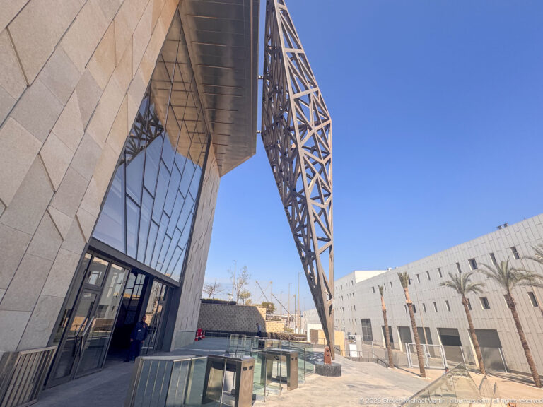 Upward exterior view of the museum’s projecting steel lattice canopy, angular stone wall, entrance glazing, stairs, and blue sky.