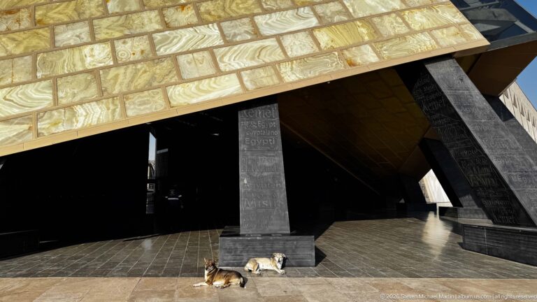 Close view beneath a gold-toned triangular museum canopy, with dark inscribed supports and two dogs resting on the stone pavement.