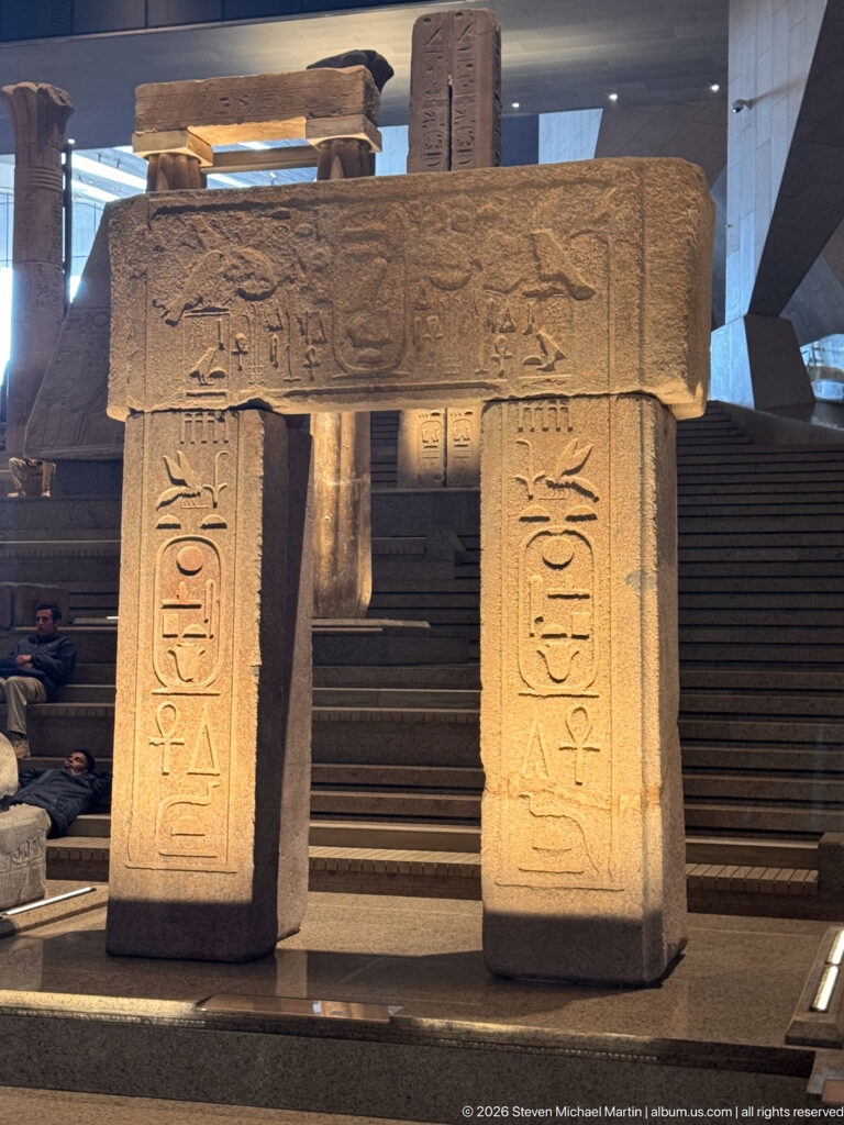 Close frontal view of an ancient Egyptian stone gateway with hieroglyphic jambs and a carved winged motif across the lintel.