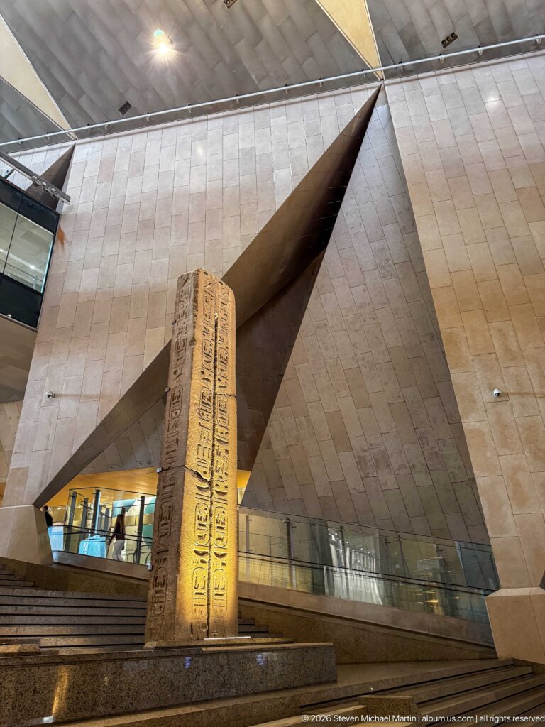Tall hieroglyph-inscribed stone block standing before angular stone walls and the Grand Egyptian Museum staircase.