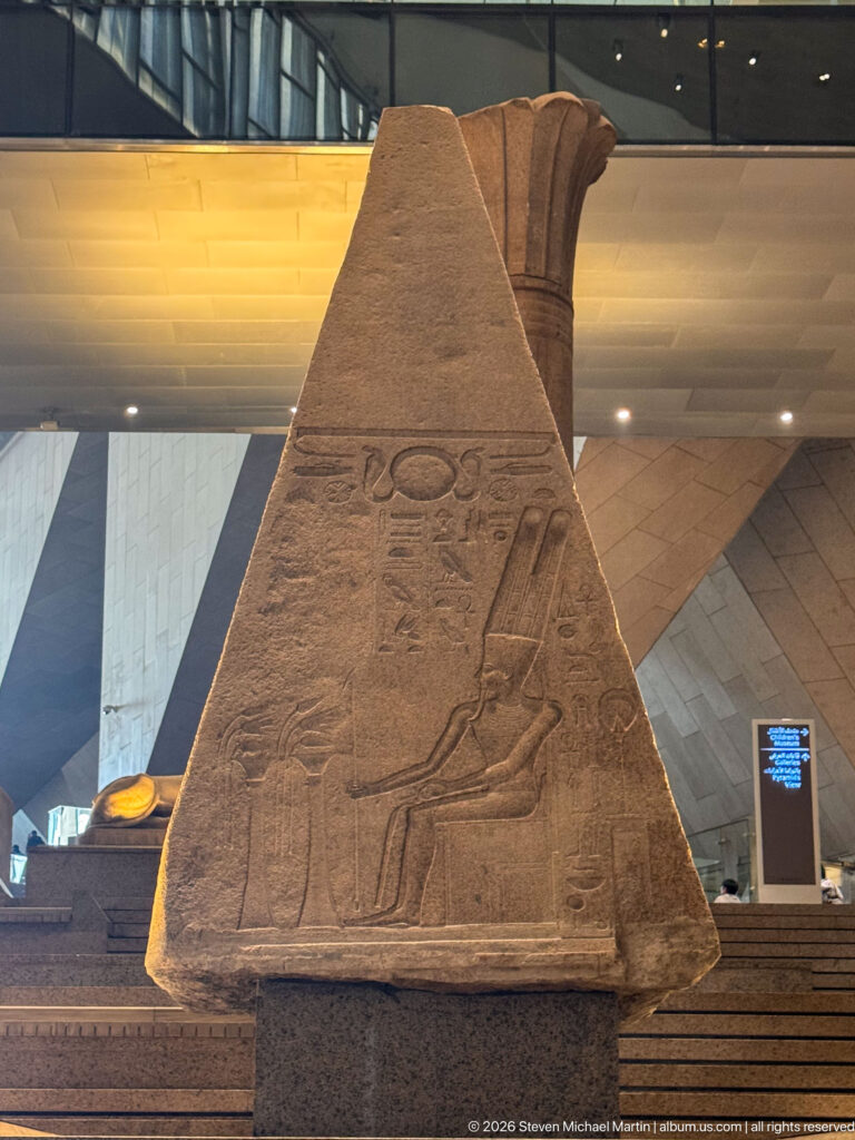 Tapered ancient stone monument with carved figures and hieroglyphs, illuminated on the Grand Egyptian Museum staircase.