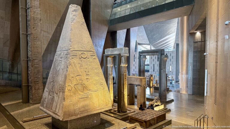 Tapered relief-carved stone monument displayed among architectural fragments beside the Grand Staircase.