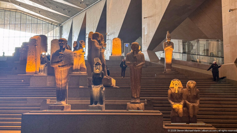 Standing and seated ancient Egyptian statues arranged on stepped platforms along the Grand Egyptian Museum staircase.