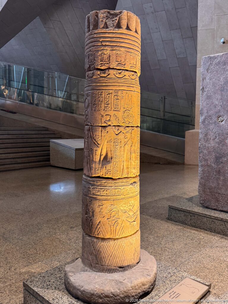 Cylindrical ancient stone column section carved with horizontal bands of figures, symbols, and hieroglyphs.