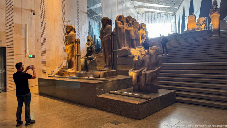 Visitor photographing rows of monumental Egyptian statues ascending the broad Grand Egyptian Museum staircase.
