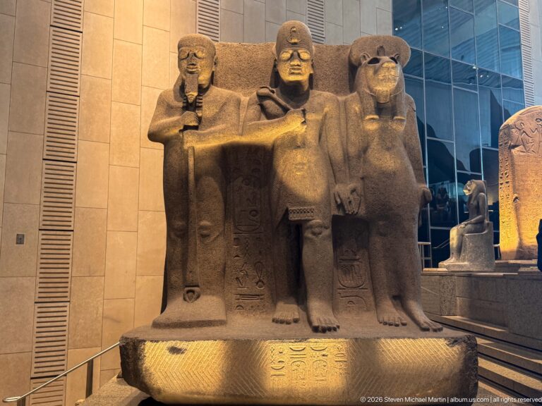 Monumental stone group of three seated Egyptian figures, with a larger central figure flanked by two companions.
