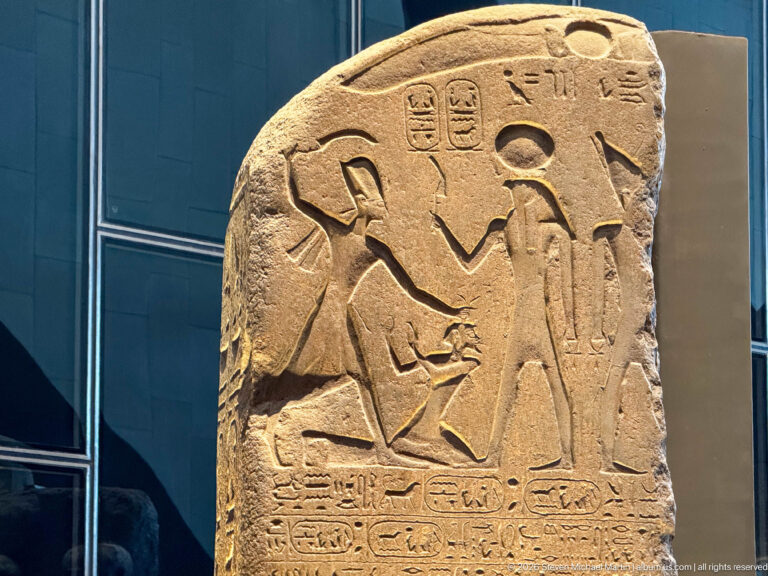 Fragmentary carved stone stela with profile figures, ritual scene, solar symbol, and horizontal hieroglyphic lines.
