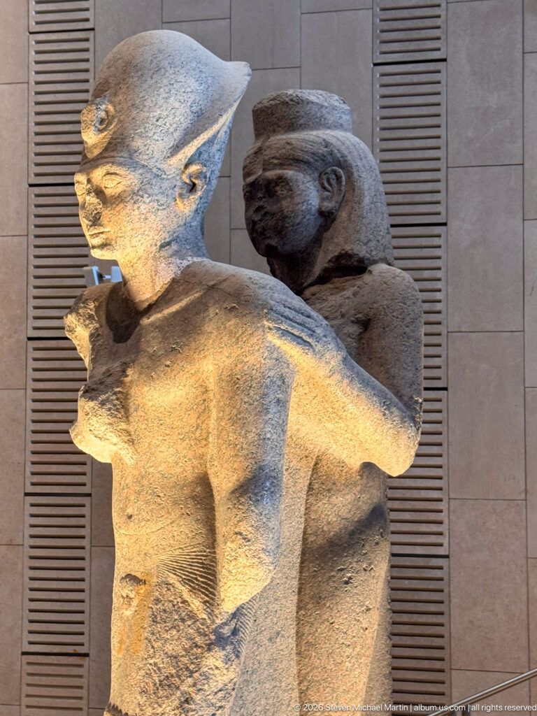 Side view of two closely joined male stone figures facing opposite directions, with broken arms and weathered surfaces.