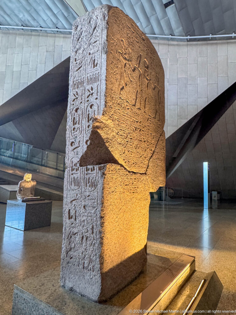 Oblique view of a massive broken stone block with hieroglyphic columns on one face and relief figures on another.