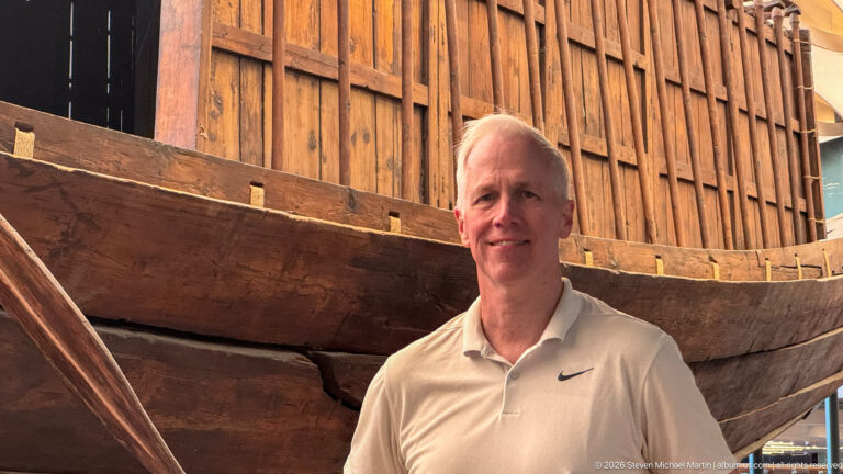 Visitor beside the tall wooden hull of King Khufu's First Boat inside the Grand Egyptian Museum's Khufu's Boats Museum.