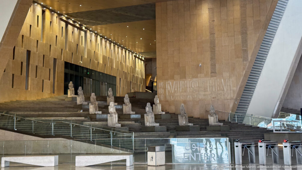 Broad Grand Staircase lined with ancient Egyptian statues on stepped platforms, with a modern escalator rising at right.