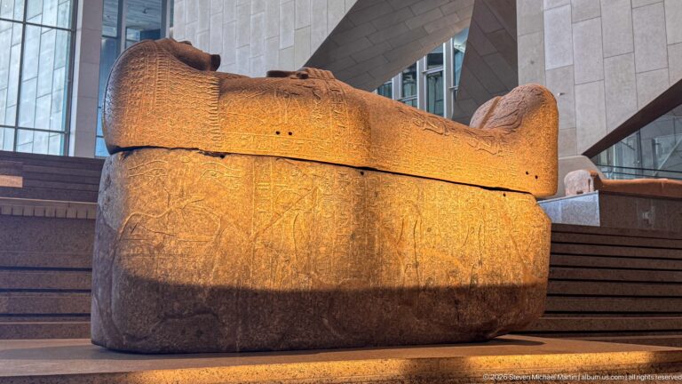 Large lidded stone sarcophagus with rounded end and worn carved decoration, illuminated beside the museum staircase.