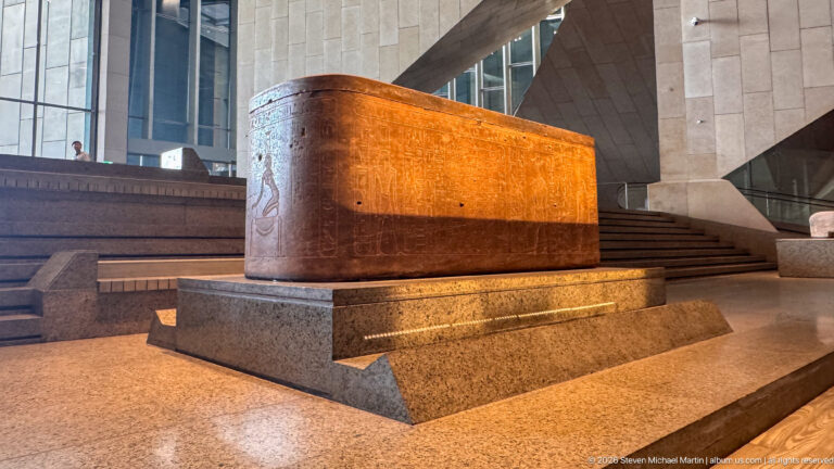 Long rectangular reddish stone sarcophagus on a raised plinth, with shallow carving near its rounded end.