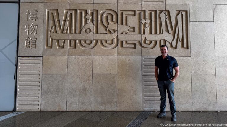 Visitor standing beneath large multilingual MUSEUM lettering mounted on a pale stone interior wall.