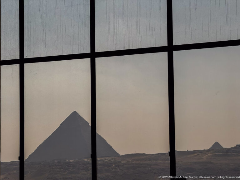 Large Giza pyramid silhouetted through dark window mullions, with a smaller pyramid visible near the right horizon.