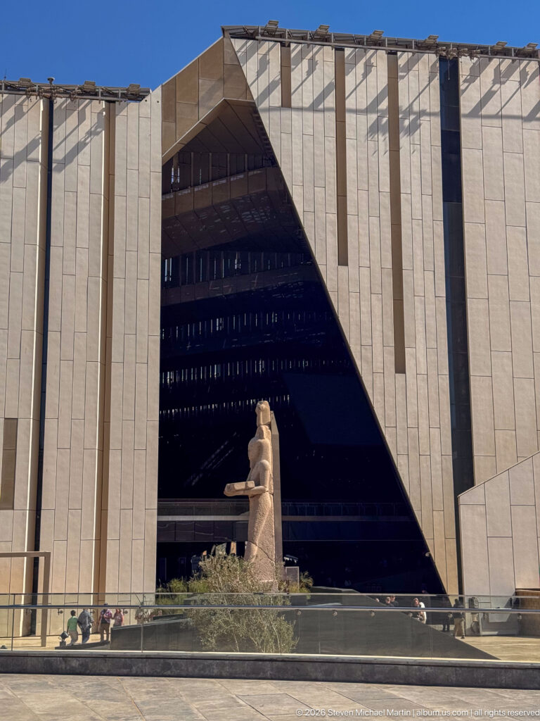 Monumental Egyptian statue centered before a huge dark triangular opening in the pale Grand Egyptian Museum façade.