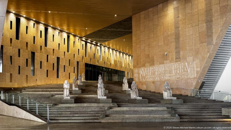 Wide interior view of the Grand Staircase with rows of ancient statues beneath a warm-lit wall and sloping structural plane.