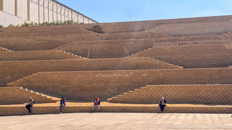 Wide stepped terraces of tan masonry with diagonal stair runs and a few seated visitors under blue sky.