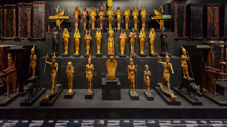 Frontal museum display of gilded shabtis, standing deities, animal figures, staffs, and dark coffin-like objects arranged symmetrically.