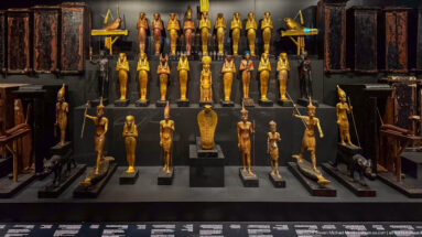 Frontal museum display of gilded shabtis, standing deities, animal figures, staffs, and dark coffin-like objects arranged symmetrically.