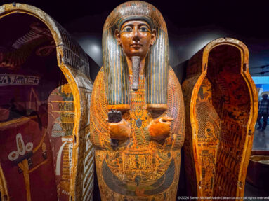 Full painted anthropoid coffin standing upright beside decorated coffin elements in a museum case.