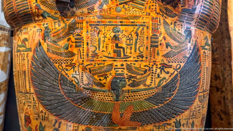 Painted coffin detail with a central winged goddess, spread feathers, hieroglyphs, and surrounding funerary figures.