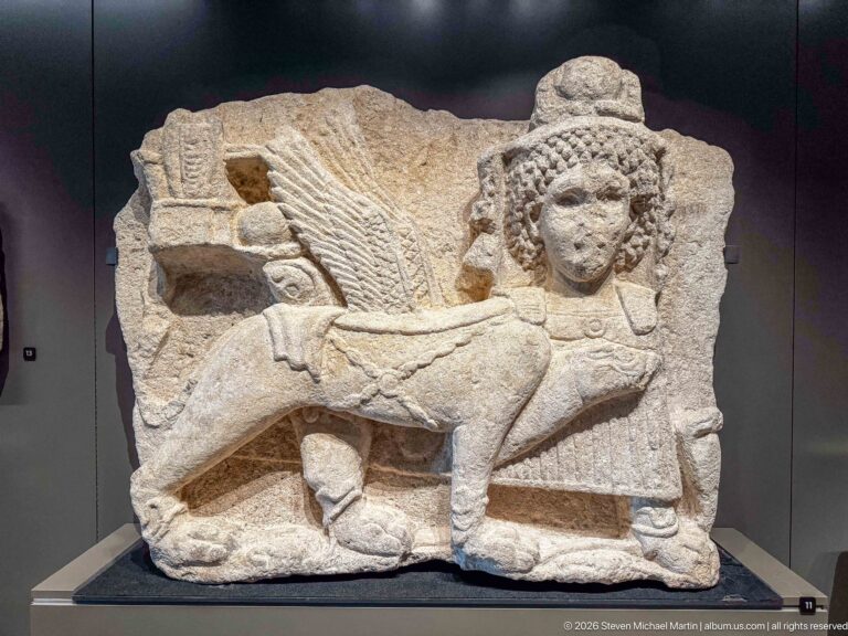 Fragmentary limestone relief with a human figure in profile, textured hair, draped clothing, and a bent leg.