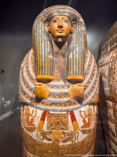 Painted anthropoid coffin with striped wig, broad beaded collar, and protective funerary figures across the chest.