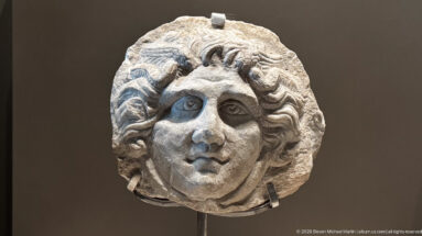 Carved stone head fragment with large eyes, rounded face, and thick curling hair.