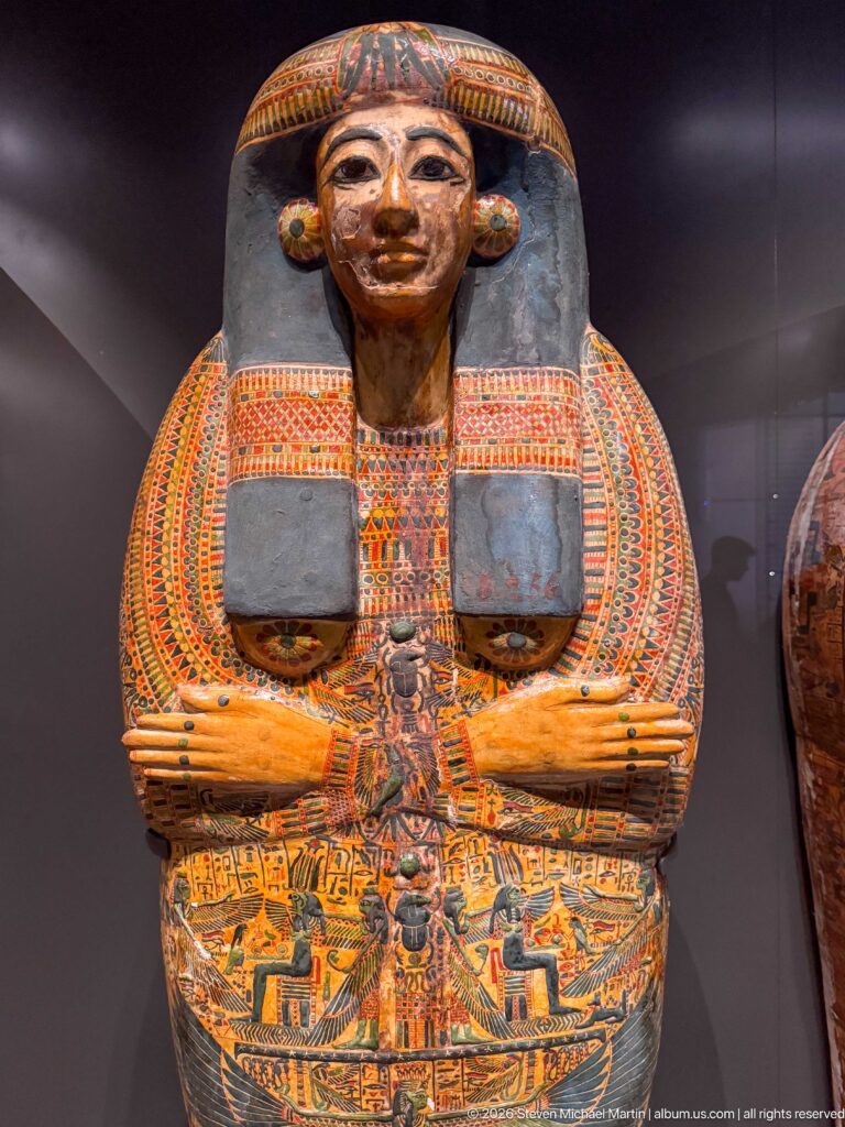 Colorfully painted anthropoid coffin with idealized face, broad collar, crossed arms, and dense funerary decoration.