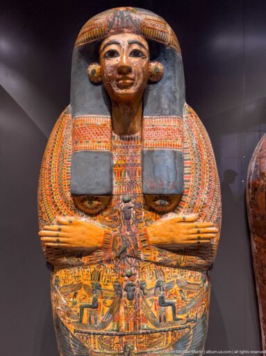 Colorfully painted anthropoid coffin with idealized face, broad collar, crossed arms, and dense funerary decoration.