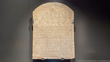 Round-topped ancient Egyptian stone stela with upper figure scene and many horizontal lines of hieroglyphs.