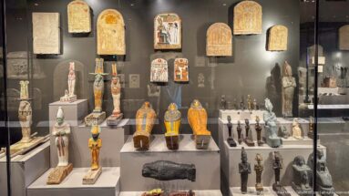 Museum case filled with ancient Egyptian coffins, stelae, funerary figures, and small ritual objects.