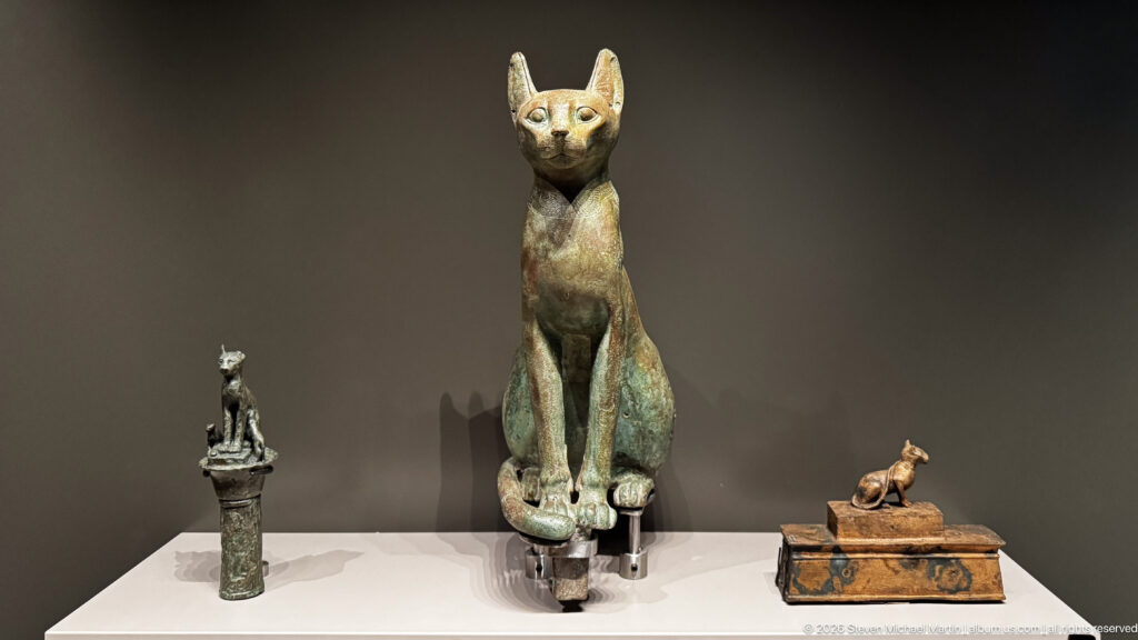 Tall seated cat statuette flanked by smaller ancient Egyptian feline figures in a museum display.