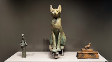 Tall seated cat statuette flanked by smaller ancient Egyptian feline figures in a museum display.