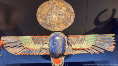 Polychrome winged scarab with blue body, spread feathered wings, red disk, and circular solar element above.