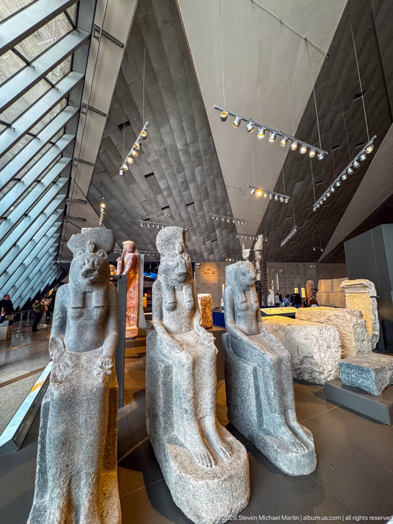 Three monumental dark-stone seated Egyptian statues displayed in a large modern gallery.