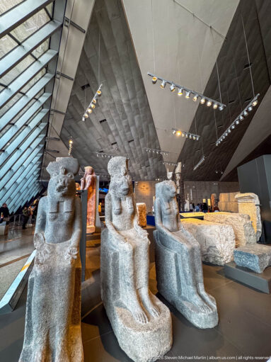 Three monumental dark-stone seated Egyptian statues displayed in a large modern gallery.