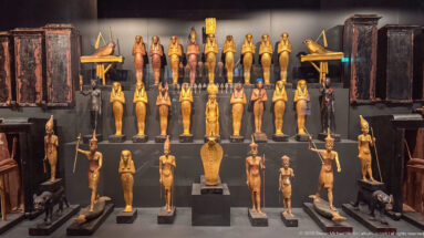 Symmetrical museum display with rows of gilded funerary figures, standing deities, dark coffin-like objects, and animal sculptures.