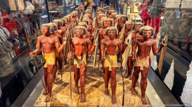 Rows of painted wooden male figures carrying long staffs or spears in a museum display case.