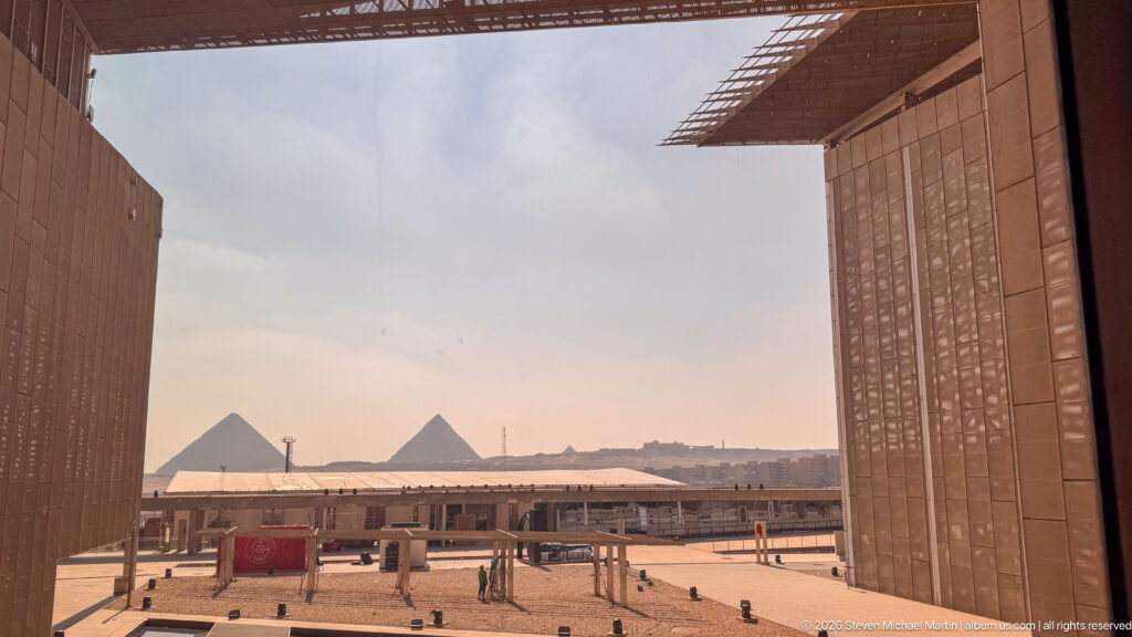 Two Giza pyramids framed between monumental museum walls above the Grand Egyptian Museum forecourt.
