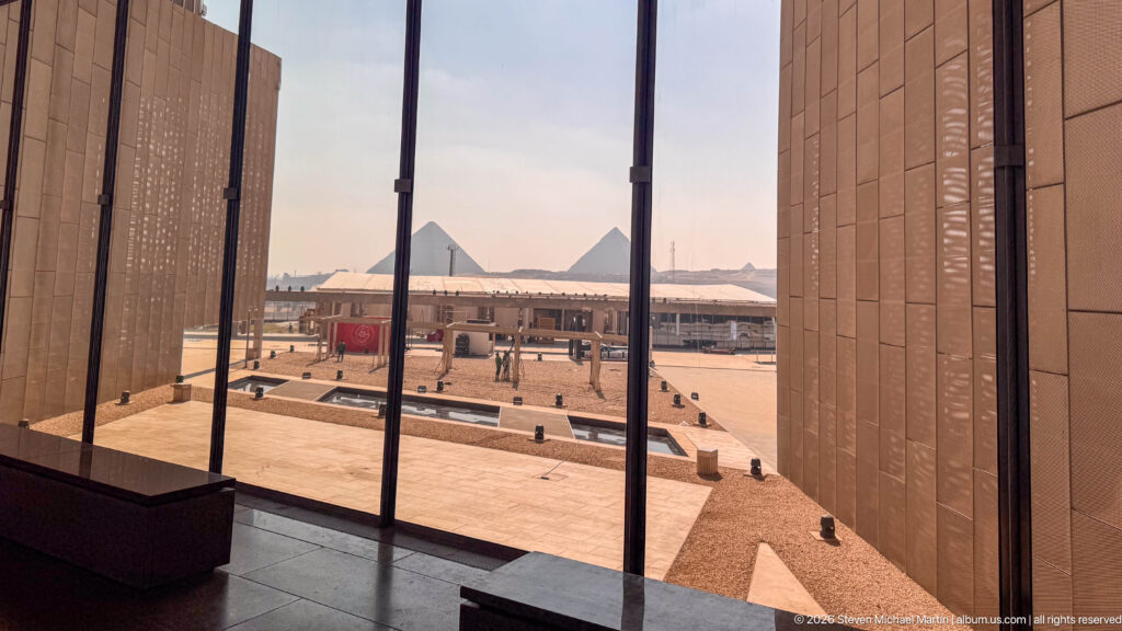 Giza pyramids seen through tall glazing from inside the Grand Egyptian Museum, beyond the museum forecourt.